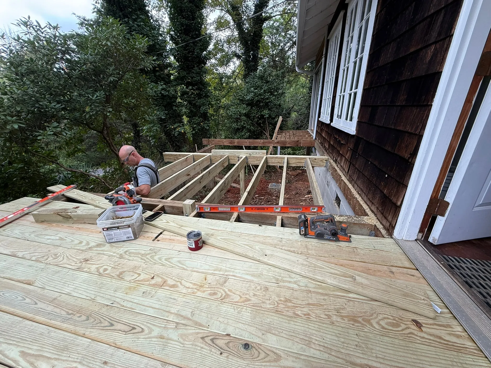 Clay Bucy cutting a joist with a circular saw while framing a new pressure-treated deck against a shingle-sided house